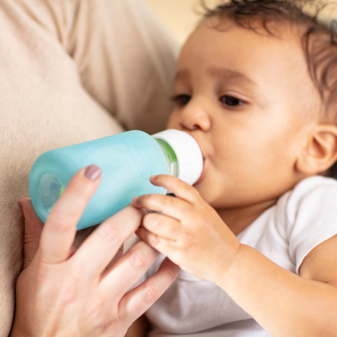 An adult holds a Dr. Brown's Natural Flow® Options+™ Narrow Glass Bottle, equipped with silicone sleeves, as a baby with short, curly hair and wearing a white shirt drinks from it.