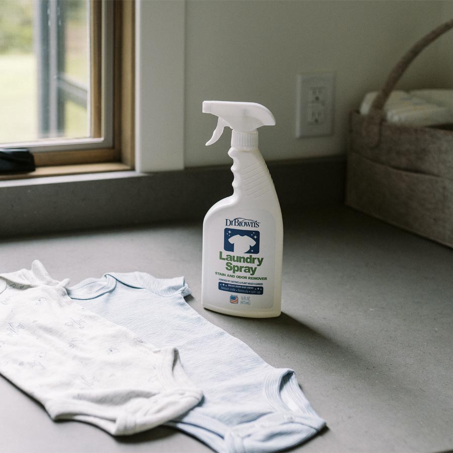 A bottle of Dr. Brown's Laundry Spray Stain and Odor Remover sits on a gray countertop next to two baby onesies. The counter, near a window with lush greenery and a basket in the background, displays this powerful formula that efficiently tackles stains and odors.