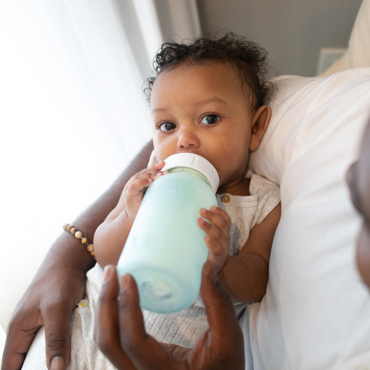 A baby nestled in someone's arms is drinking milk from Dr. Brown's Natural Flow® Options+™ Wide-Neck Glass Bottle, its silicone sleeve gleaming softly. The person gently holds the bottle as natural light streams through a window, creating a serene and tender scene.