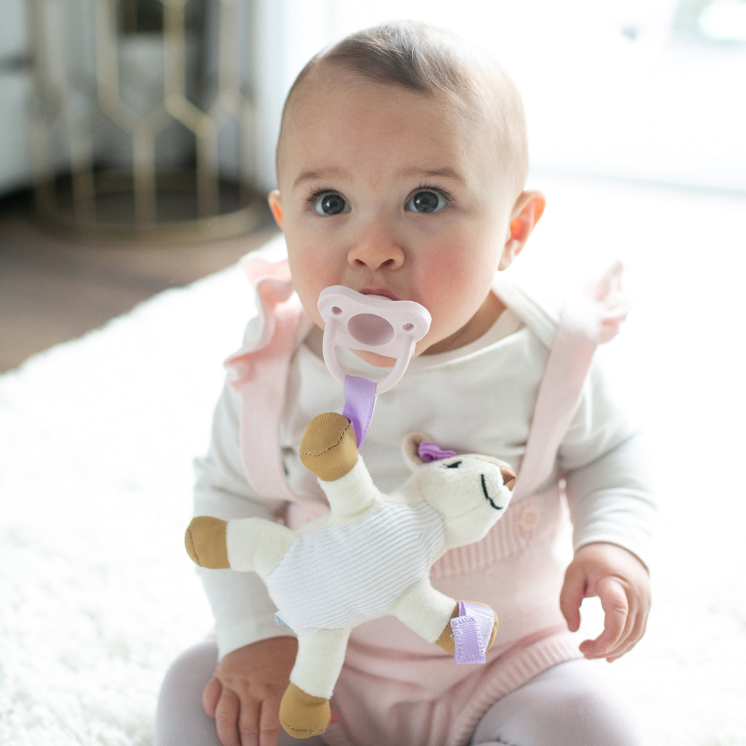 A baby sits on a soft, white rug, holding Dr. Brown's® Lovey Pacifier and Teether Holder in the Deer design. Dressed in a white outfit with pink overalls, they are surrounded by warmth and comfort as a decorative metal stand graces the background.