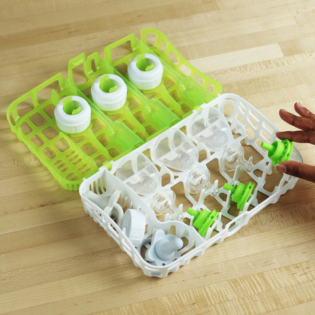 A Dr. Brown’s™ Options+™ Dishwasher Basket for Baby Bottle Parts in green and white is open on a wooden surface. It neatly holds several baby bottle components, such as nipples and lids. A hand extends toward the basket from the right side of the image.