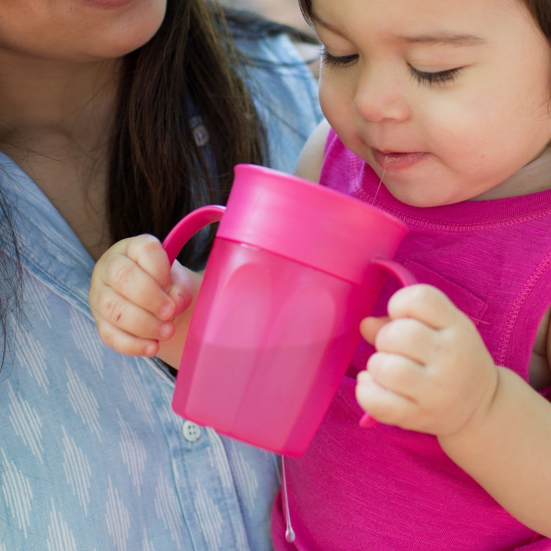 Wearing a pink shirt, a toddler drinks from the Dr. Brown’s® Milestones™ Cheers360™ Cup with Handles, 7 oz/200 mL, as some liquid spills out unexpectedly. An adult in partial view observes with curiosity while the child experiments with this 360-degree cup in a delightful outdoor close-up scene.