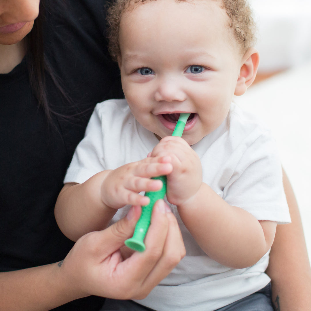 A baby with curly hair and bright eyes is holding Dr. Brown’s™ Toddler Toothbrush, Crocodile, 1-Pack with super-soft bristles, smiling. An adult gently guides the toothbrush. The background is softly blurred, focusing on the child's joyful expression and the toothbrush's eye-catching design by Dr. Brown's.