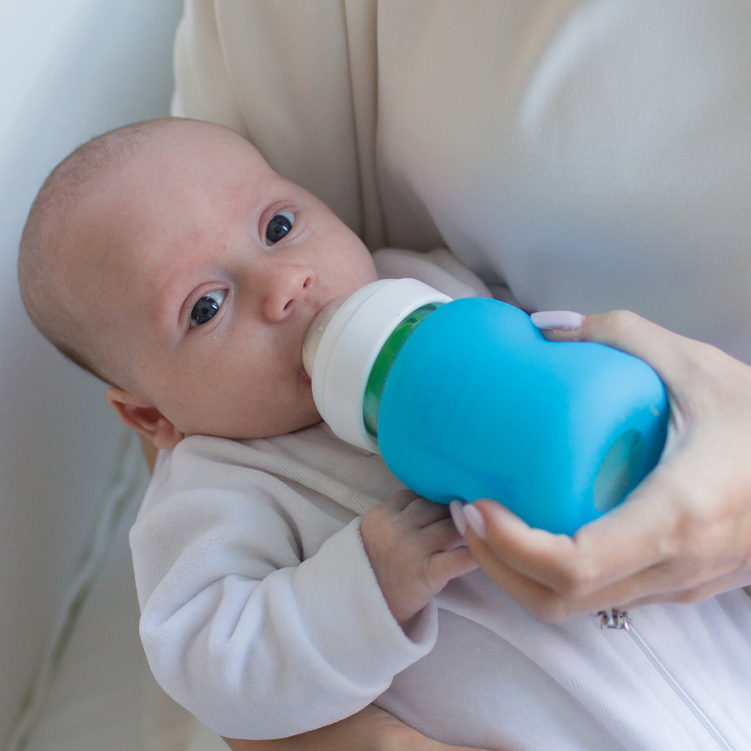 A baby in a white outfit is being fed with a blue baby bottle, nestled in someone's arms on a white cushion. The bottle is equipped with Dr. Brown's Natural Flow® Options+™ Wide-Neck Glass Bottle Silicone Sleeves, providing both style and grip with its white and blue design.