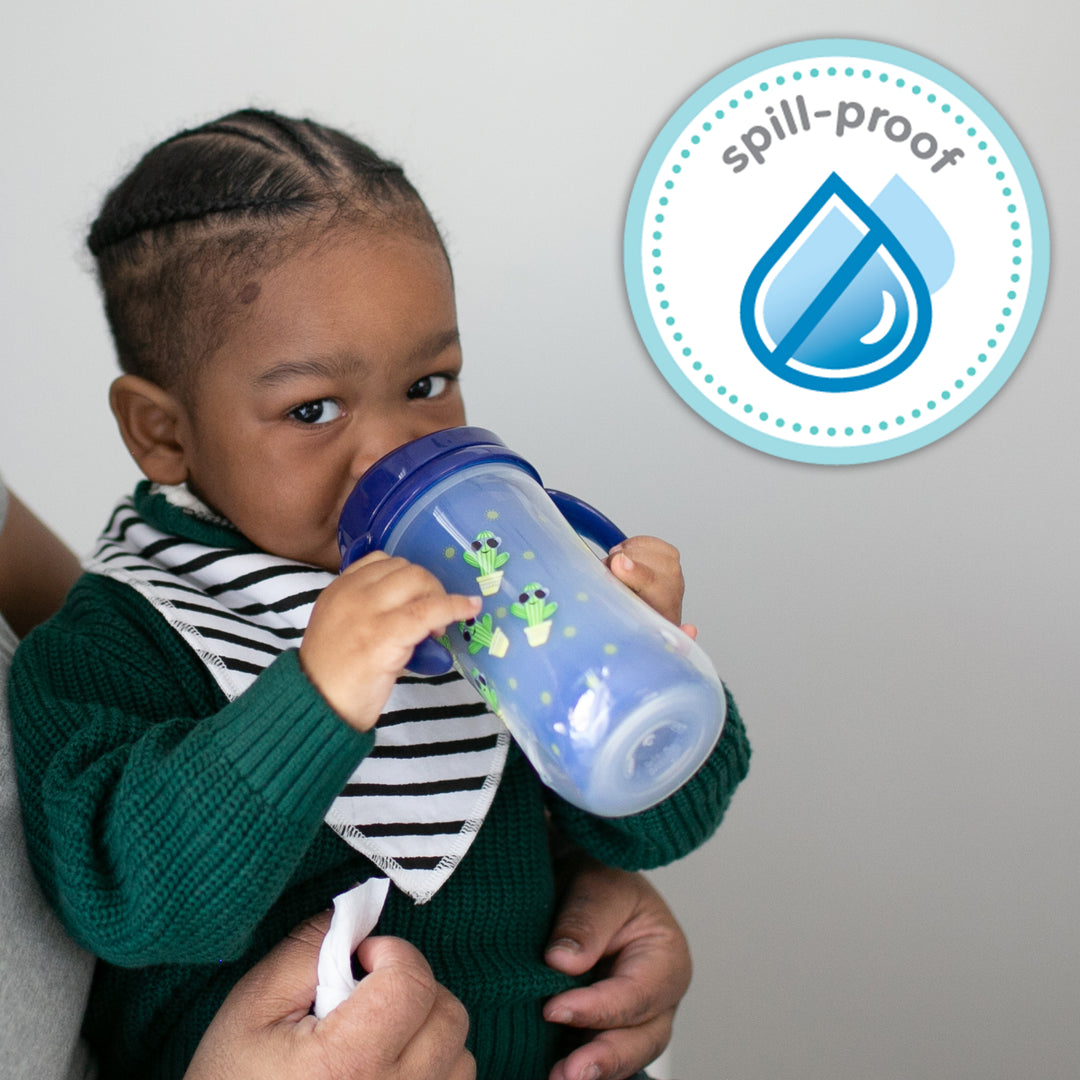 A toddler sips from a blue Dr. Brown's Milestones™ Insulated Hard Spout Sippy Cup, featuring green frog designs, while being supported by an adult. In the top right corner, a "spill-proof" icon with two blue droplets highlights the cup's durable hard spout design.