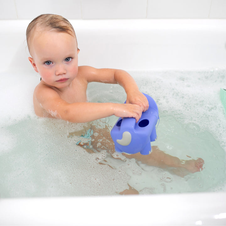 In a bubble-filled bathtub with white tiled walls, a toddler with wet hair sits focused on enjoying bath time fun while boosting motor skills development. The child holds Dr. Brown’s™ CleanUp™ Pour & Roar Watering Can Bath Toy alongside a purple elephant-shaped toy.