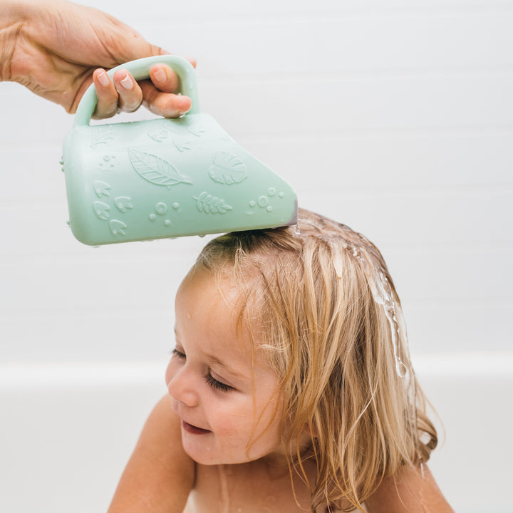 A young child with blonde hair is being rinsed with water from Dr. Brown’s™ CleanUp™ Dino-Pour Baby Bath Rinse Cup. The child is sitting in a bathtub, smiling with eyes closed, enjoying the tear-free bath time as the water gently flows down their hair.