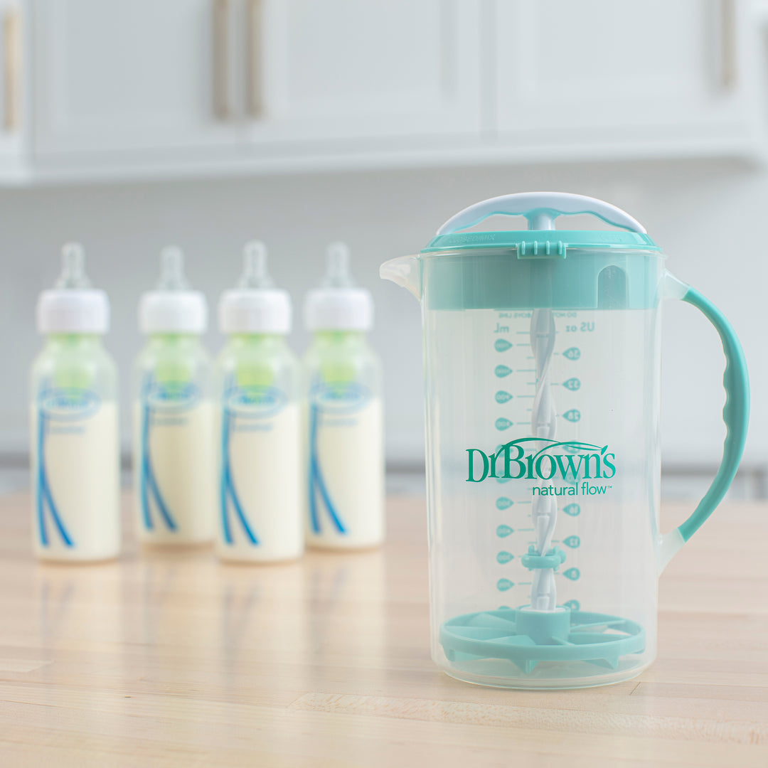 On the kitchen counter sits a Dr. Brown's Natural Flow® Formula Mixing Pitcher, showcasing its transparent design with a teal lid and no-drip spout. In the background, four baby bottles are filled with milk, prepared for feeding time.