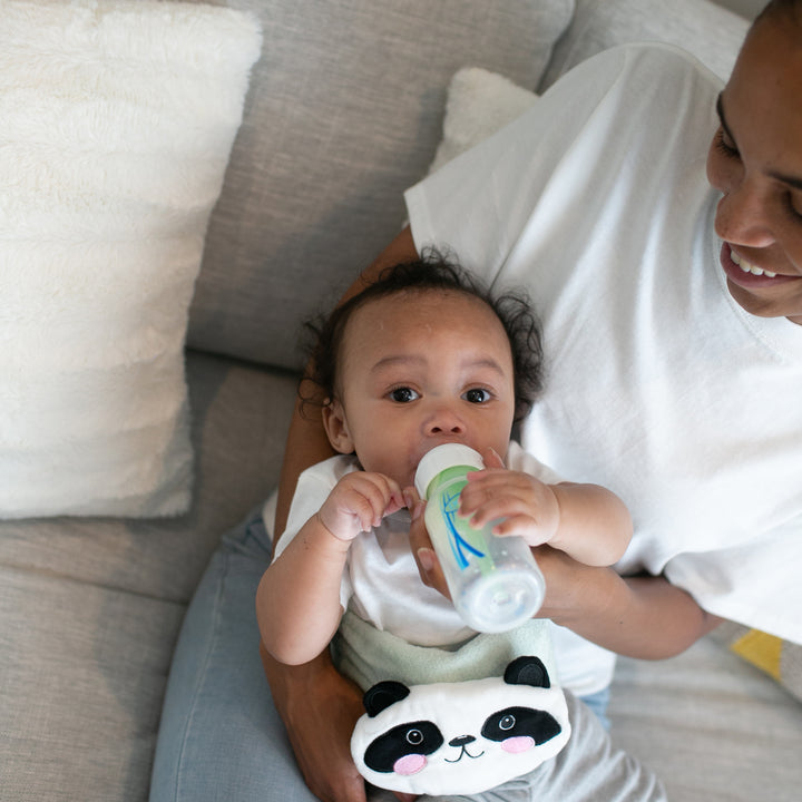 Settled in an adult's lap on a light-colored couch, a baby drinks from a bottle, wrapped in warmth and comfort. Dressed in a white panda-themed outfit, this serene moment echoes the soothing effects of Dr. Brown’s™ Infant Gripebelt™ Colic Relief Belt. The smiling adult complements the scene with a crisp white shirt.