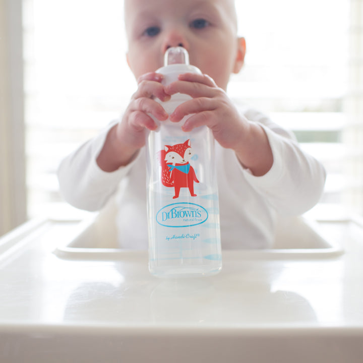 A baby sits in a high chair, holding a Dr. Brown's® Milestones™ Narrow Sippy Bottle that boasts an illustration of a red fox. The bottle is equipped with a soft silicone spout, ideal for transitioning from bottle to cup. The softly blurred background draws attention to the child's endearing grip.