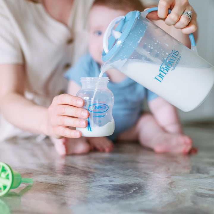 A person fills a baby's bottle with milk from Dr. Brown's Natural Flow® Formula Mixing Pitcher by Dr. Brown's, which is designed to reduce gas. In the background, a partially visible baby in a light blue outfit sits on the marble surface, with a green pacifier lying nearby.