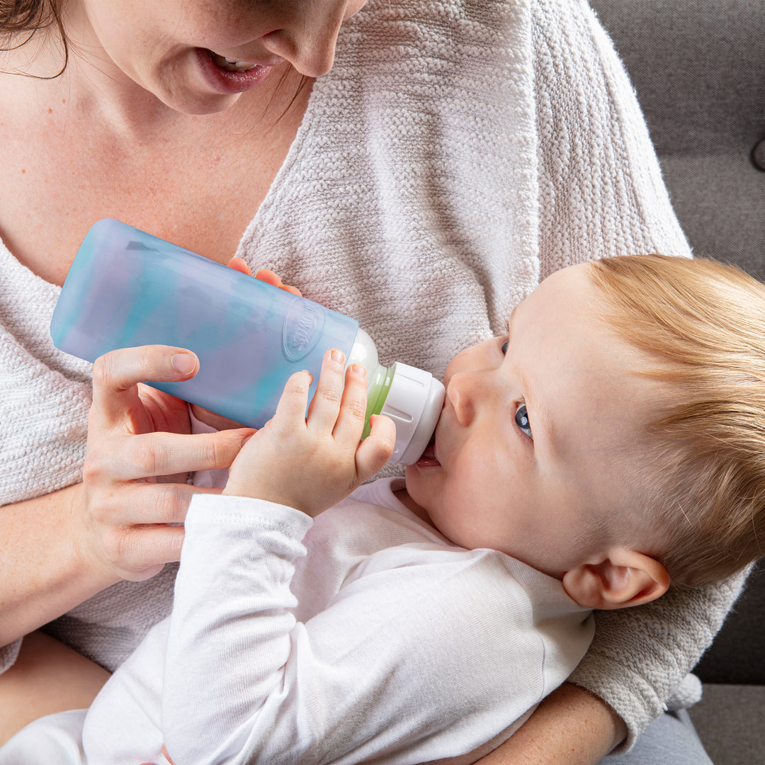 A baby in a white outfit is being cradled by an adult wearing a light-colored sweater, while drinking from Dr. Brown's Natural Flow® Options+™ Narrow Glass Bottle. The focused infant holds onto the protective silicone sleeve provided with the bottle during their meal.