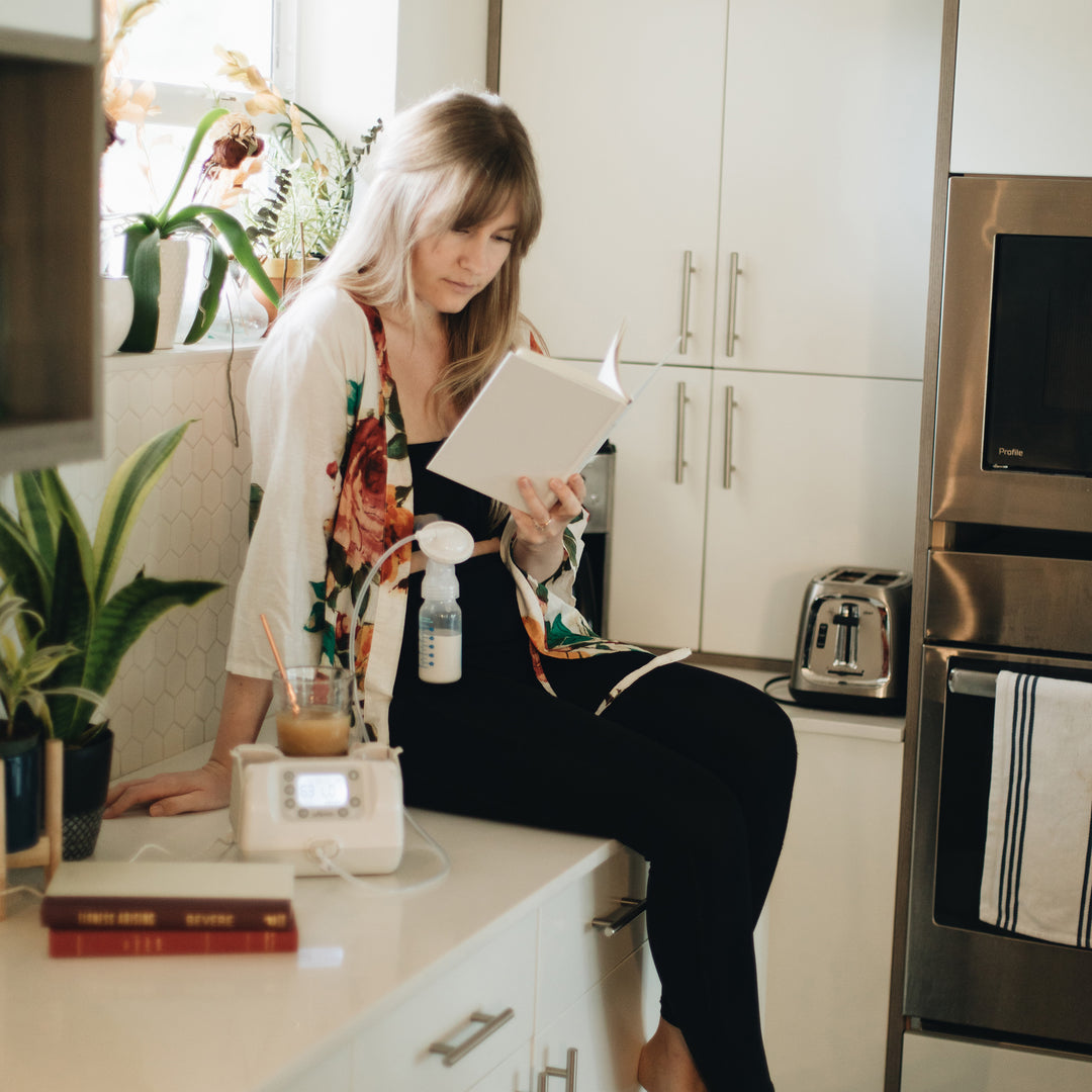 A woman sits on a kitchen counter, absorbed in her book while wearing a floral cardigan. She utilizes Dr. Brown's Hands Free Pumping Bra with an electric pump. The backdrop features various kitchen appliances and plants, accompanied by a nearby glass of iced coffee.