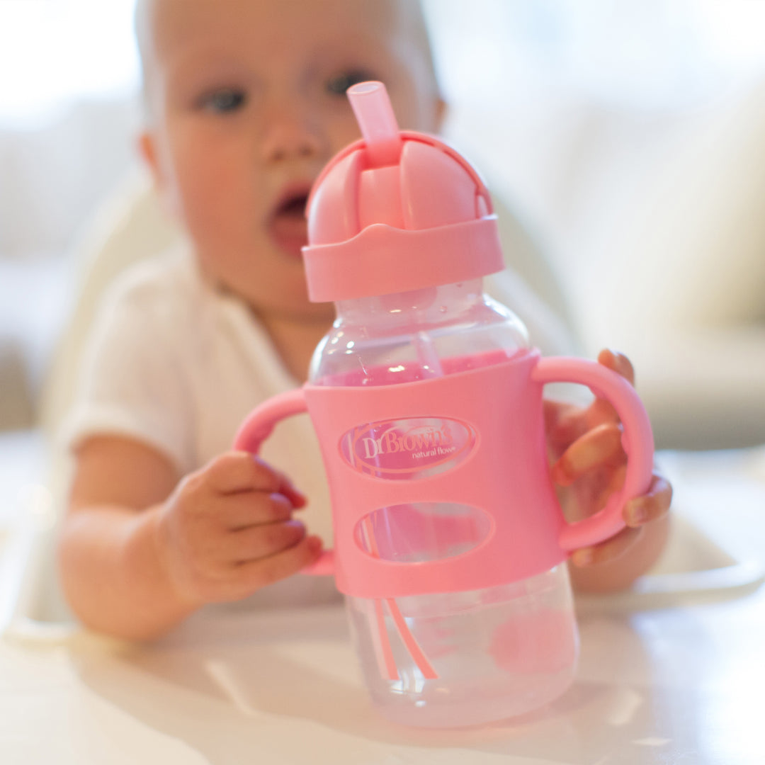 A baby in a high chair is holding Dr. Brown’s™ Milestones™ Wide-Neck Sippy Straw Bottle, featuring silicone handles and a weighted straw, in pink. The baby seems intent on honing independent drinking skills, set against a softly blurred background that creates a cozy, homey atmosphere.