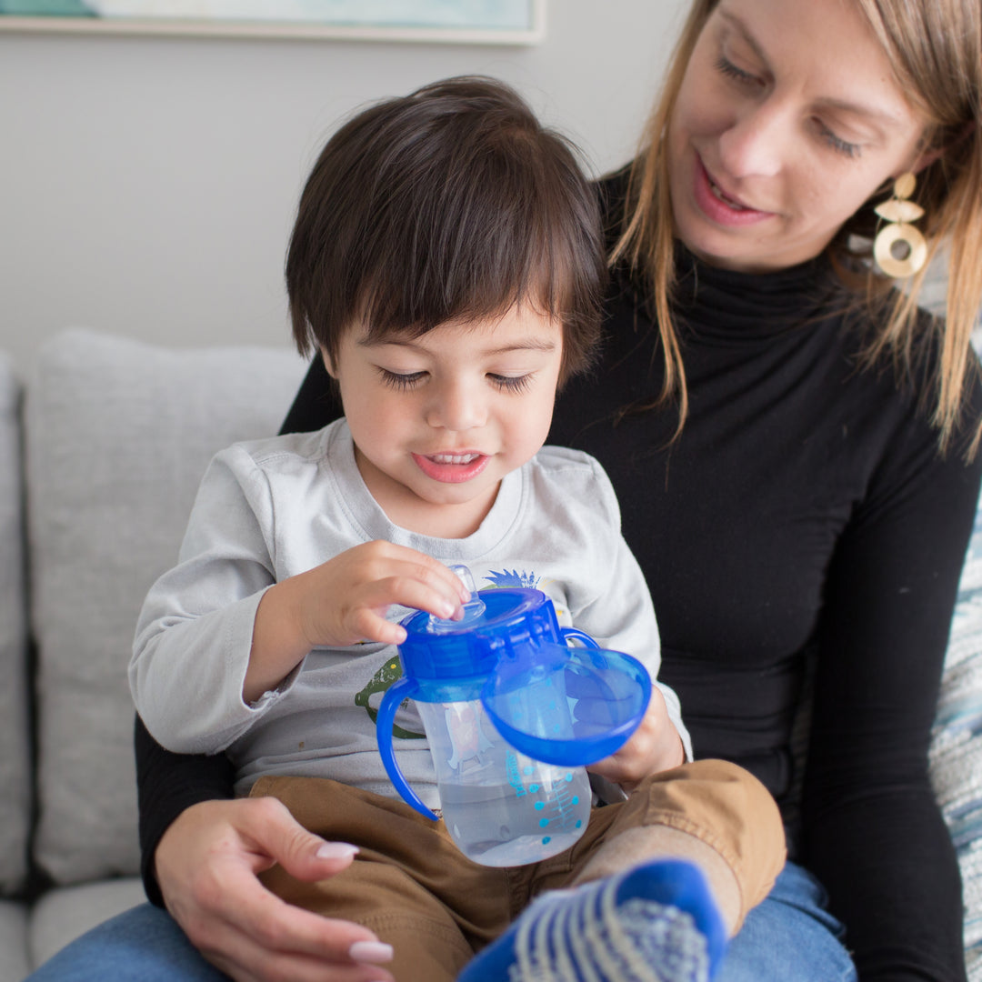 A woman with light brown hair holds a toddler on her lap, who is absorbed in a Dr. Brown’s® Soft-Spout Toddler Cup, 9 oz/270 ml (9m+), designed for spill-free learning. The child, dressed in a long-sleeved gray shirt, smoothly navigates the transition from bottle to cup as they both sit comfortably on a light-colored couch.