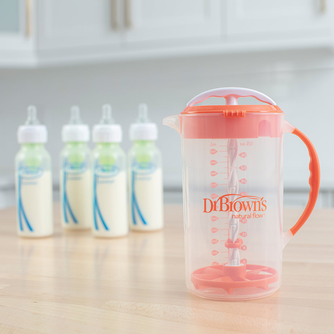 A Dr. Brown's Natural Flow® Formula Mixing Pitcher, featuring an orange handle and mixing mechanism, is positioned on the kitchen counter. In the background, four baby bottles filled with milk stand in a row, ready to help reduce gas. The softly blurred kitchen perfectly accentuates the pitcher and bottles.