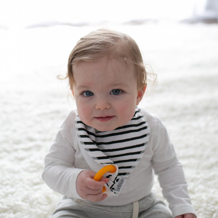 A baby with light brown hair sits on a soft white surface, wearing a white long-sleeve shirt and Dr. Brown’s™ Bandana Bibs with Removable Teether. Holding an orange item and looking toward the camera, this adorable scene is both charming and practical thanks to the bib's removable teether feature from Dr. Brown's.