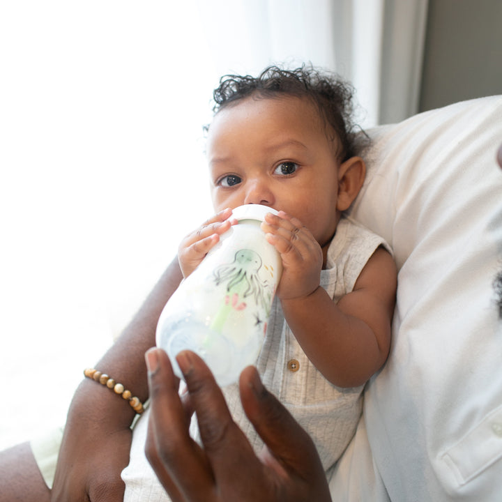 A curly-haired baby is drinking from a Dr. Brown’s Natural Flow® Anti-Colic Options+™ Wide-Neck Baby Bottle, adorned with an octopus design. An adult in a white shirt with a beaded bracelet is holding the baby, as soft natural light filters through a nearby window.