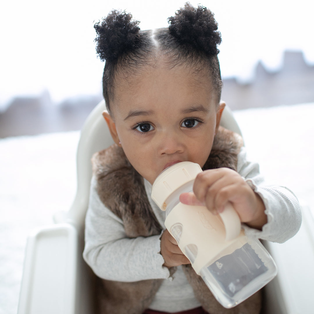 A toddler with curly hair styled in two buns sits in a high chair, mastering drinking skills with the Dr. Brown's® Milestones™ Wide-Neck Sippy Spout Bottle, which features silicone handles. The child wears a fluffy vest over a long-sleeve top, as the softly blurred background completes the scene.