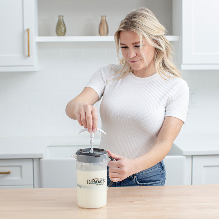 A person in a white shirt uses the Dr. Brown's Natural Flow® Formula Mixing Pitcher on a kitchen counter. They are preparing a liquid mixture, possibly to reduce gas, in a modern kitchen setting with white cabinets and decorative vases in the background.