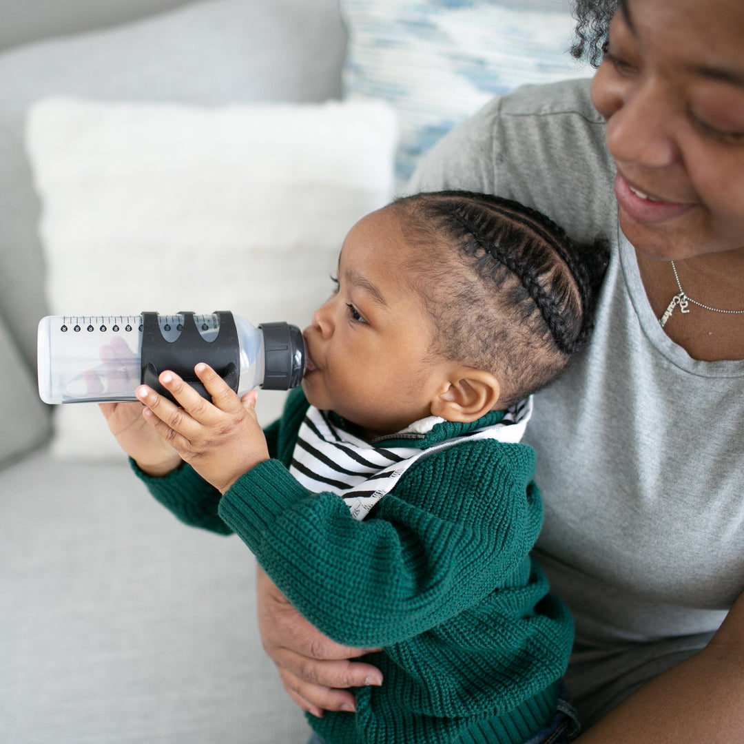 A baby with braided hair, dressed in a green sweater and striped bib, sips from a Dr. Brown’s® Milestones™ Narrow Sippy Spout Bottle with Silicone Handles while sitting on an adult's lap. The adult, wearing a gray shirt, smiles warmly at the child as they transition from bottle nipple. They are seated on a light-colored couch.