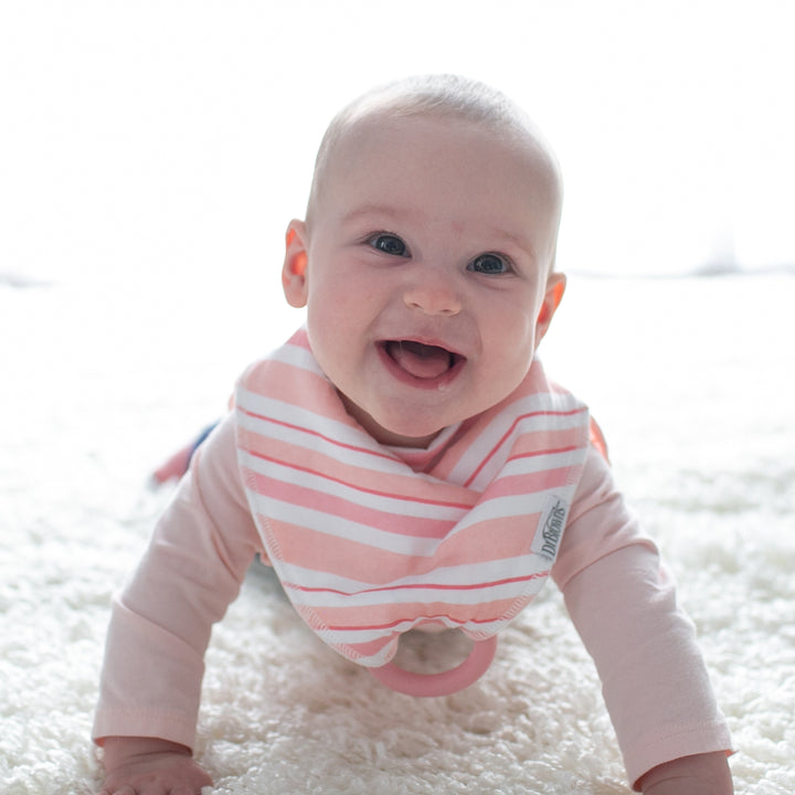 A baby is seen crawling on a pale carpet, smiling widely with visible teeth, while wearing a Dr. Brown’s™ Bandana Bib with Removable Teether in a super-absorbent pink and white design. The softly lit background accentuates the baby's joyful expression.