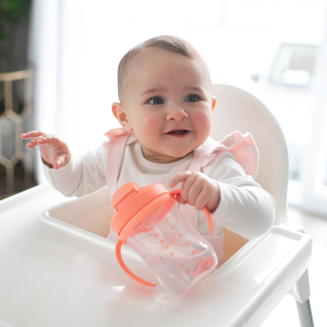 A baby sits in a high chair, smiling in a white outfit with pink accents. Clutched in their tiny hands is Dr. Brown’s™ Milestones™ Baby’s First Straw Cup, featuring a weighted straw and pink lid, perfect for the bottle-to-cup transition. The softly blurred background suggests a bright room filled with natural light.