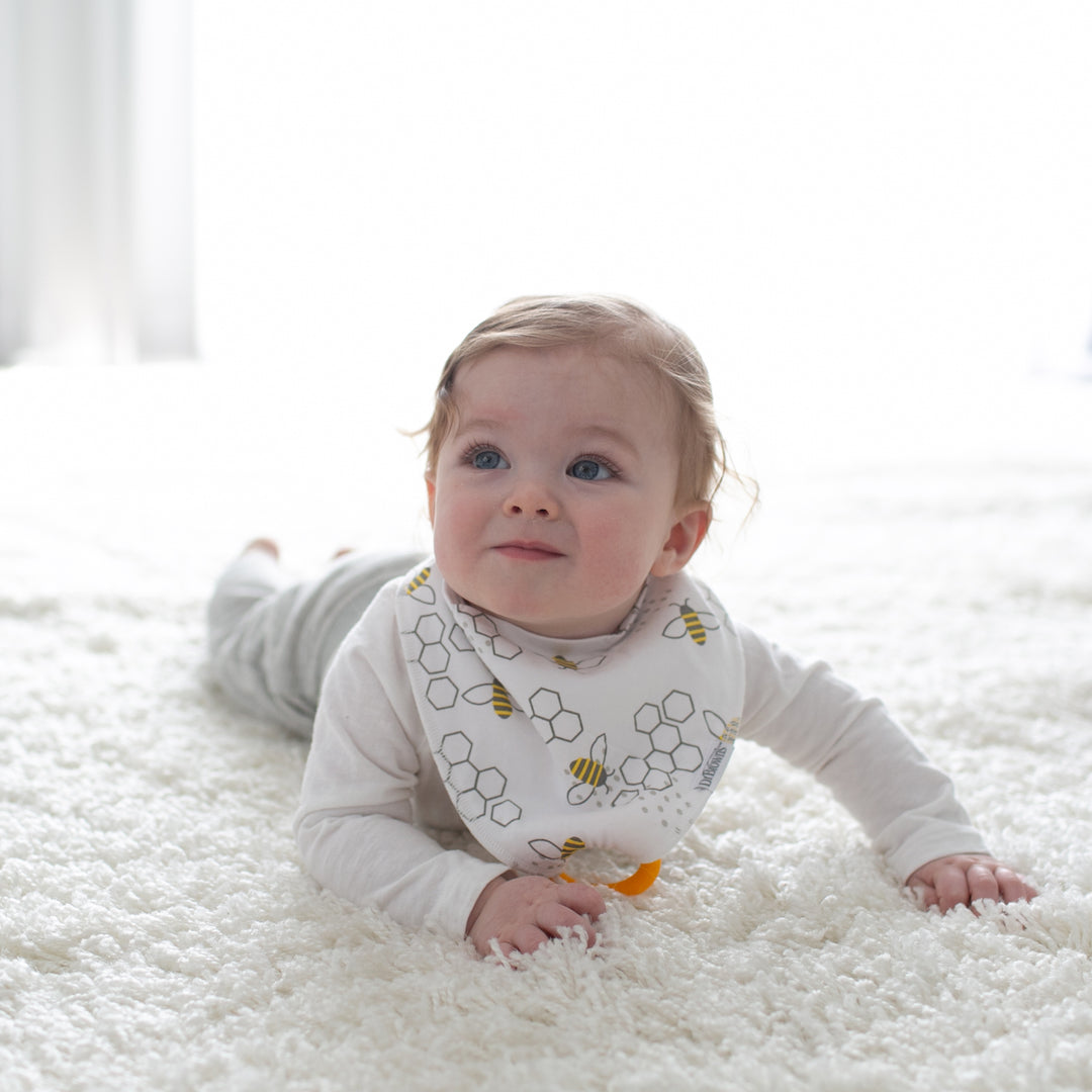 A baby with light hair and blue eyes lies on a soft white rug, dressed in a cozy white outfit and a Dr. Brown’s™ Bandana Bib with Removable Teether featuring charming bee and honeycomb patterns. The gentle lighting enhances the serene atmosphere, creating an ideal setting for peaceful play.