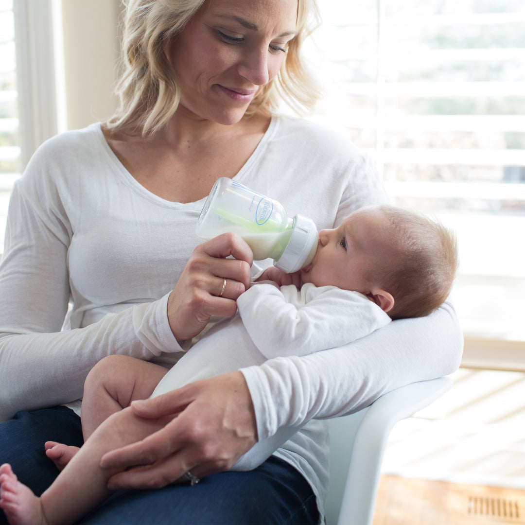 A woman in a white shirt is sitting and smiling gently as she feeds a baby with Dr. Brown’s Natural Flow® Anti-Colic Options+™ Wide-Neck Glass Baby Bottle, featuring a Level 1 Slow Flow Nipple. The baby, dressed in white, is cradled in her arm. They are sitting by a bright window where the natural flow of sunlight creates a serene atmosphere.