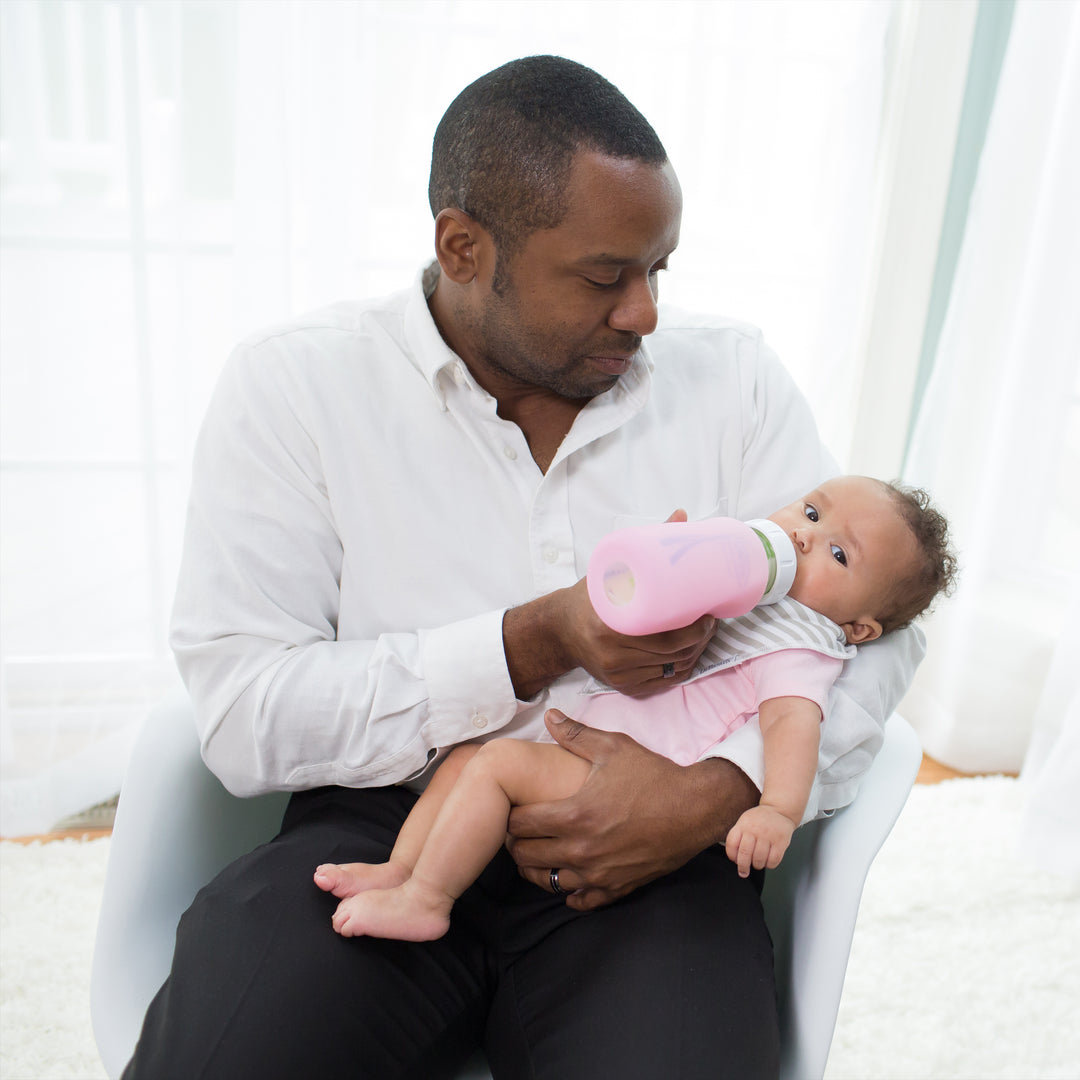 A man in a white shirt sits in a chair feeding a baby with Dr. Brown's Natural Flow® Options+™ Wide-Neck Glass Bottle Silicone Sleeve. The baby, wearing pink and gray, is cradled in his arms while the background is softly lit with sheer curtains.