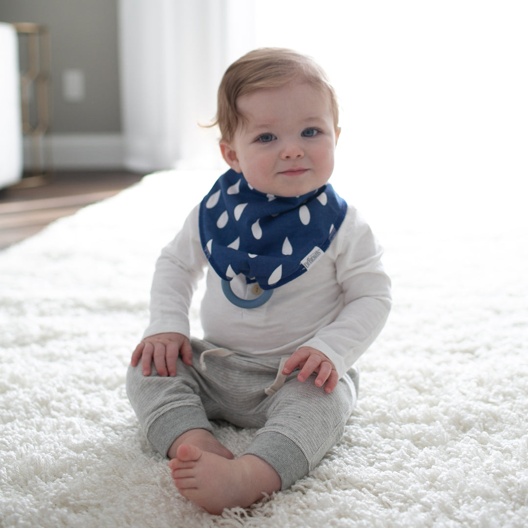 A teething baby with light brown hair sits on a fluffy white rug, dressed in gray pants and a white long-sleeve outfit. The charm of the softly lit room in the background is enhanced by a Dr. Brown’s™ Bandana Bib featuring white raindrop patterns and an attached removable teether.