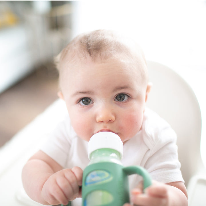 A baby with light hair and blue eyes is sitting in a high chair, holding onto Dr. Brown's Dr. Brown’s® Milestones™ Narrow Silicone Handles as they practice independent drinking skills with their bottle. The softly blurred background emphasizes the baby's focused gaze during this transition from bottle to cup.