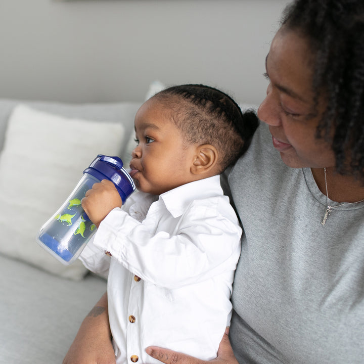 A caregiver holds a toddler on a couch. The child, dressed in a white shirt, drinks from Dr. Brown’s Milestones™ Insulated Straw Cup. The adult, wearing a gray shirt, gazes affectionately at the child while a pillow rests in the background.