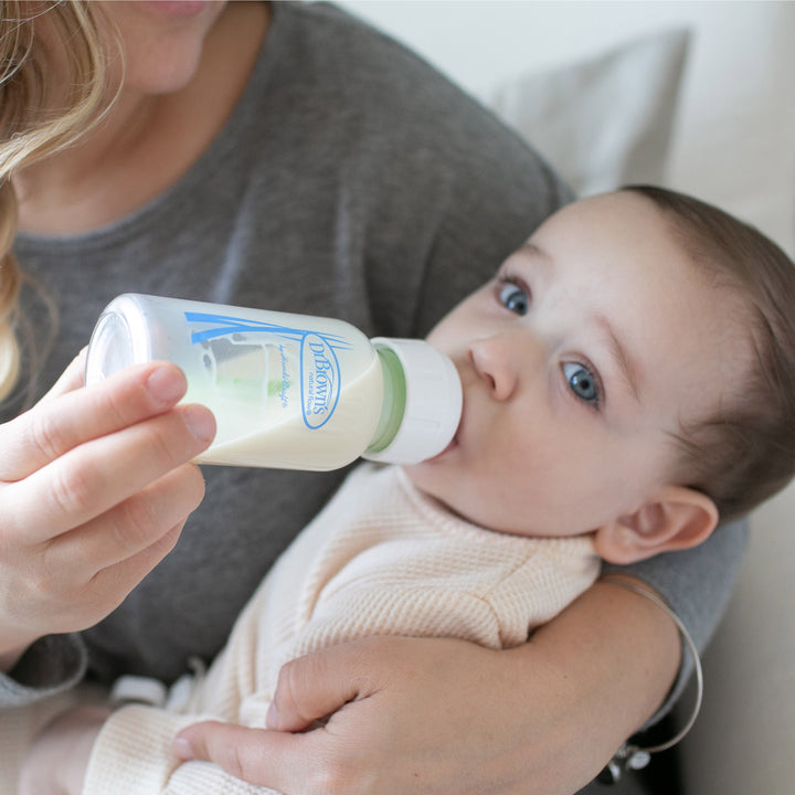 A light-haired, blue-eyed baby is drinking from Dr. Brown's Natural Flow® Anti-Colic Options+™ narrow glass baby bottle with silicone sleeve while being held by an adult in a gray top. The bottle features a breast-like nipple with branded text, creating a soft-lit and cozy setting ideal for feeding time.