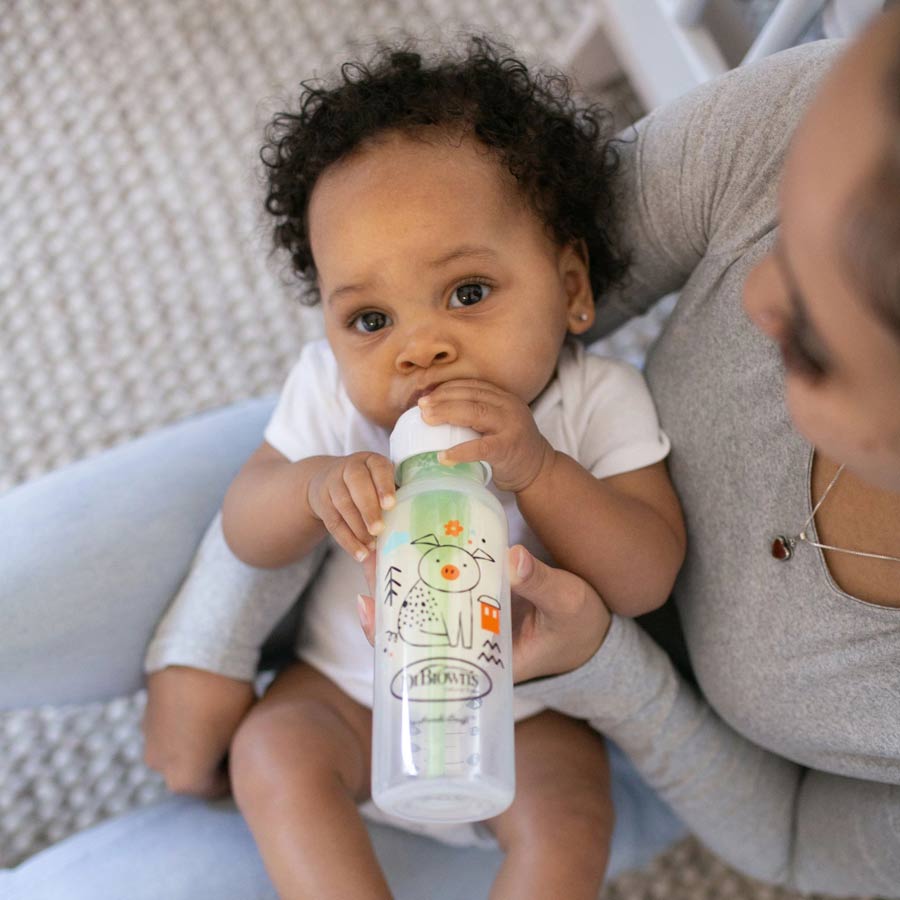 A baby with curly hair is drinking from a Dr. Brown’s Natural Flow® Anti-Colic Options+™ Narrow Baby Bottle, which features a whimsical animal design. The infant is held by an adult wearing a gray top, sitting on a textured, light-colored surface.