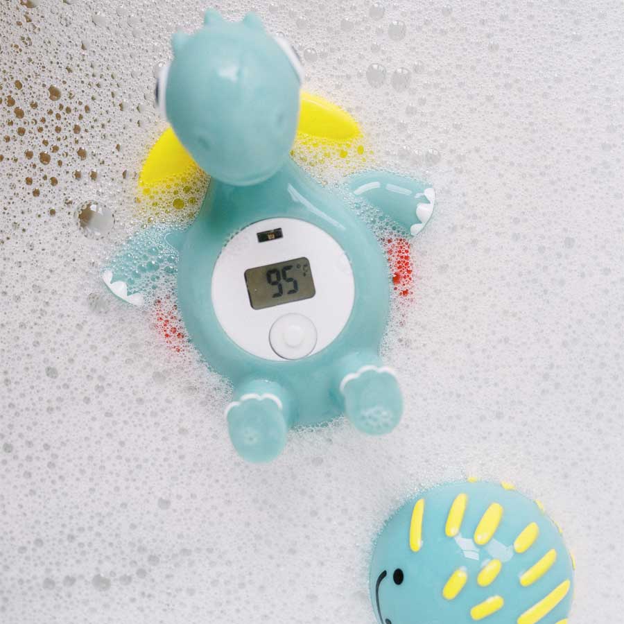 A Dr. Brown's™ CleanUp™ Temposaurus™ Floating Bath Thermometer, shaped like a light blue dinosaur, bobs in the bubble bath, its digital screen showing a water temperature of 95°F. Nearby, a smiling yellow and blue bath toy peeks through the suds.