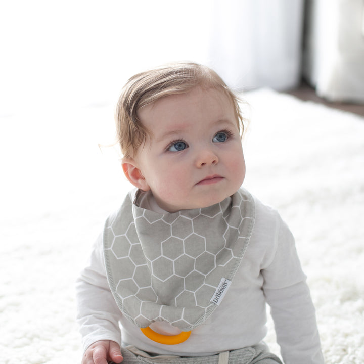 A baby with light brown hair and blue eyes sits on a soft, white rug. They are wearing a white shirt and Dr. Brown's™ Bandana Bib with Removable Teether, featuring a geometric pattern in grey, while holding the yellow teether attachment. The background is softly lit.