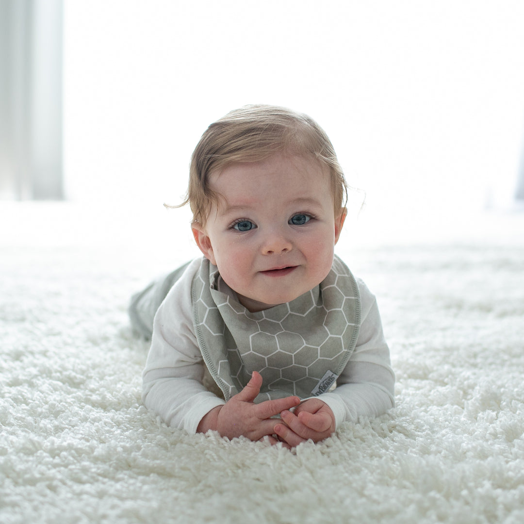 A baby wearing Dr. Brown’s™ Bandana Bibs with a removable teether in a gray pattern is lying on a fluffy white carpet, smiling as soft, natural light enhances the scene. The super-absorbent fabric from Dr. Brown's ensures comfort, while the detachable teether adds a playful touch to this charming moment.