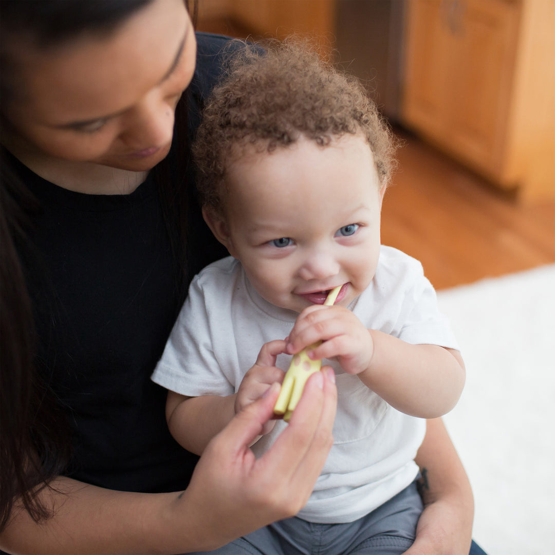 A caregiver dressed in a black shirt gently assists a joyful toddler with curly hair, who is wearing a white shirt, in using the Dr. Brown’s™ Infant-to-Toddler Toothbrush featuring a giraffe design. The toddler giggles while concentrating on their oral hygiene routine, as they sit together on the light-colored floor indoors.