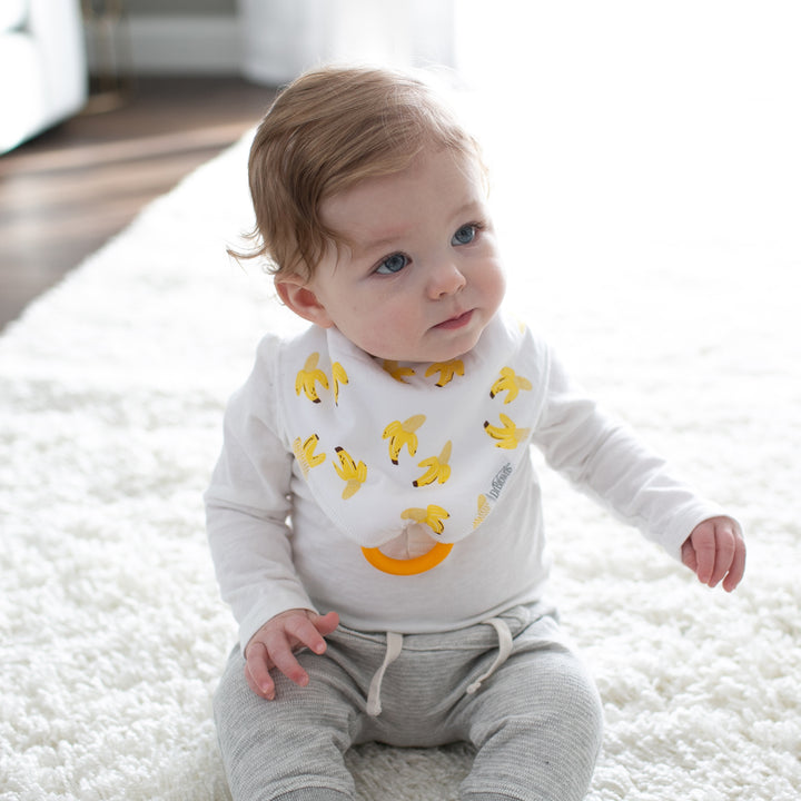 A baby with light brown hair sits on a white fuzzy carpet, wearing a white long-sleeve shirt, gray pants, and a Dr. Brown's Bandana Bibs adorned with banana patterns featuring a removable teether. The room is bright and softly lit.