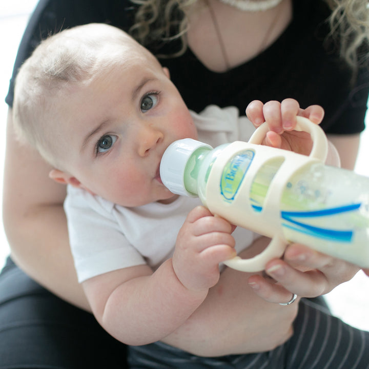 A baby with short hair is independently drinking from a Dr. Brown’s bottle, equipped with Dr. Brown’s® Milestones™ Narrow Silicone Handles for easy grip. The baby, dressed in a white shirt, sits comfortably on an adult's lap against a softly blurred background.