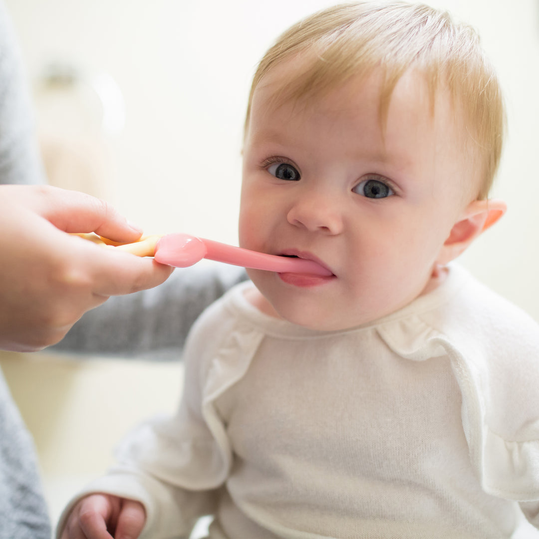 A baby in a white shirt sits while an adult feeds them with a pink spoon, their curious eyes matching the short light brown hair. Nearby, a Dr. Brown’s™ Flamingo Toddler Toothbrush with super-soft bristles and a suction-cup base patiently waits as the background gently blurs, highlighting this tender moment.