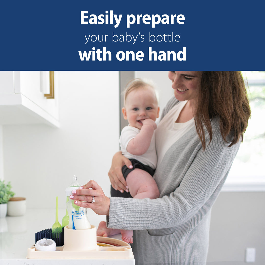 In the kitchen, a woman balances her baby on her hip as she utilizes the bottle prep station. The text above states, "Effortlessly prepare your baby's bottle with one hand." Several baby bottles and Dr. Brown’s™ 2-in-1 Dry & Prep Silicone Mat from Dr. Brown's are neatly arranged on the counter.