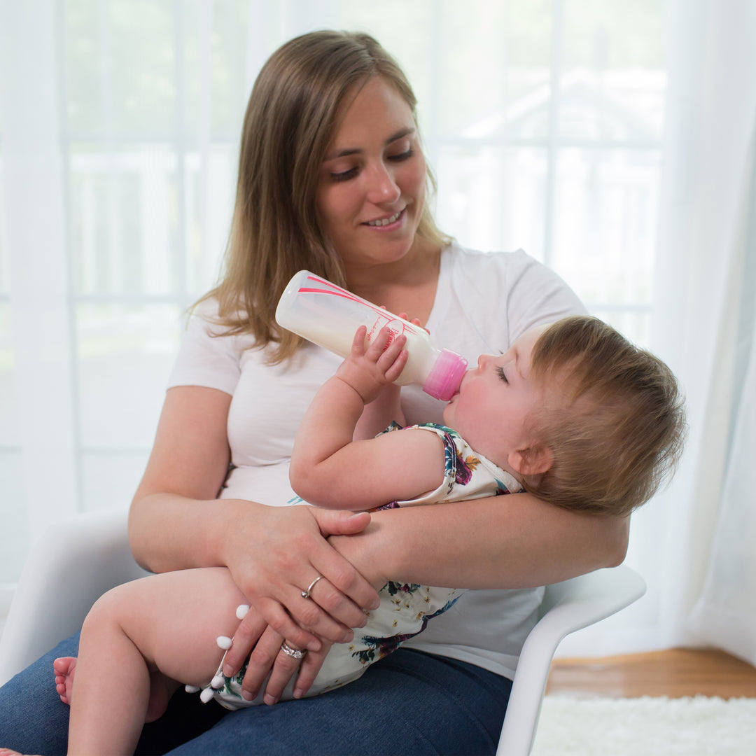 A woman sitting on a chair holds a baby in her lap, who is drinking from a Dr. Brown's Natural Flow® Anti-Colic Options+™ Narrow Baby Bottle, featuring a Level 1 Slow Flow Nipple. The woman smiles warmly at the baby in the brightly lit room with sheer curtains providing an elegant backdrop.