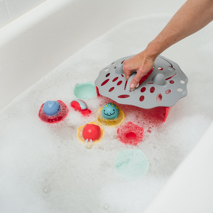 A person is using a gray Dr. Brown’s™ CleanUp™ Dino-Scoop Baby Bath Toy Organizer to collect vibrant bath toys, including red, blue, and smiley face toys, from the bubbly bathtub. The playful scene with white foam surrounding the toys is made even more convenient by the handy organizer from Dr. Brown's.