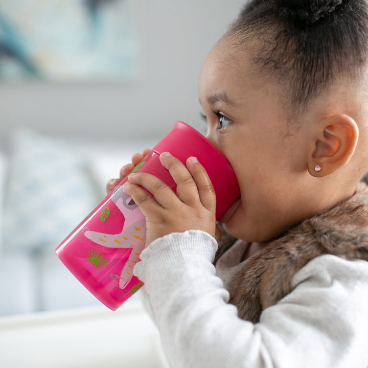 A toddler with curly hair sips from a pink Dr. Brown's® Milestones™ Cheers360™ Cup, adorned with animal designs. The child is dressed in a light-colored top and a fur vest. The softly blurred background reveals an elegantly furnished room in light tones, ideal for mess-free learning.