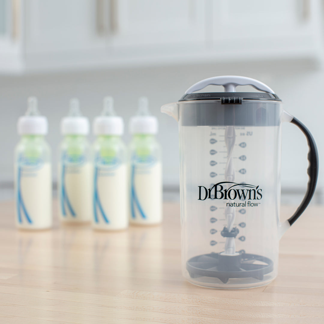 A Dr. Brown's Natural Flow® Formula Mixing Pitcher with measurement markings and a convenient handle is placed on a kitchen counter. Behind it, four baby bottles containing milk, each equipped with a nipple and cap designed to minimize gas for infants, are lined up. Blurred kitchen cabinets can be seen in the background.