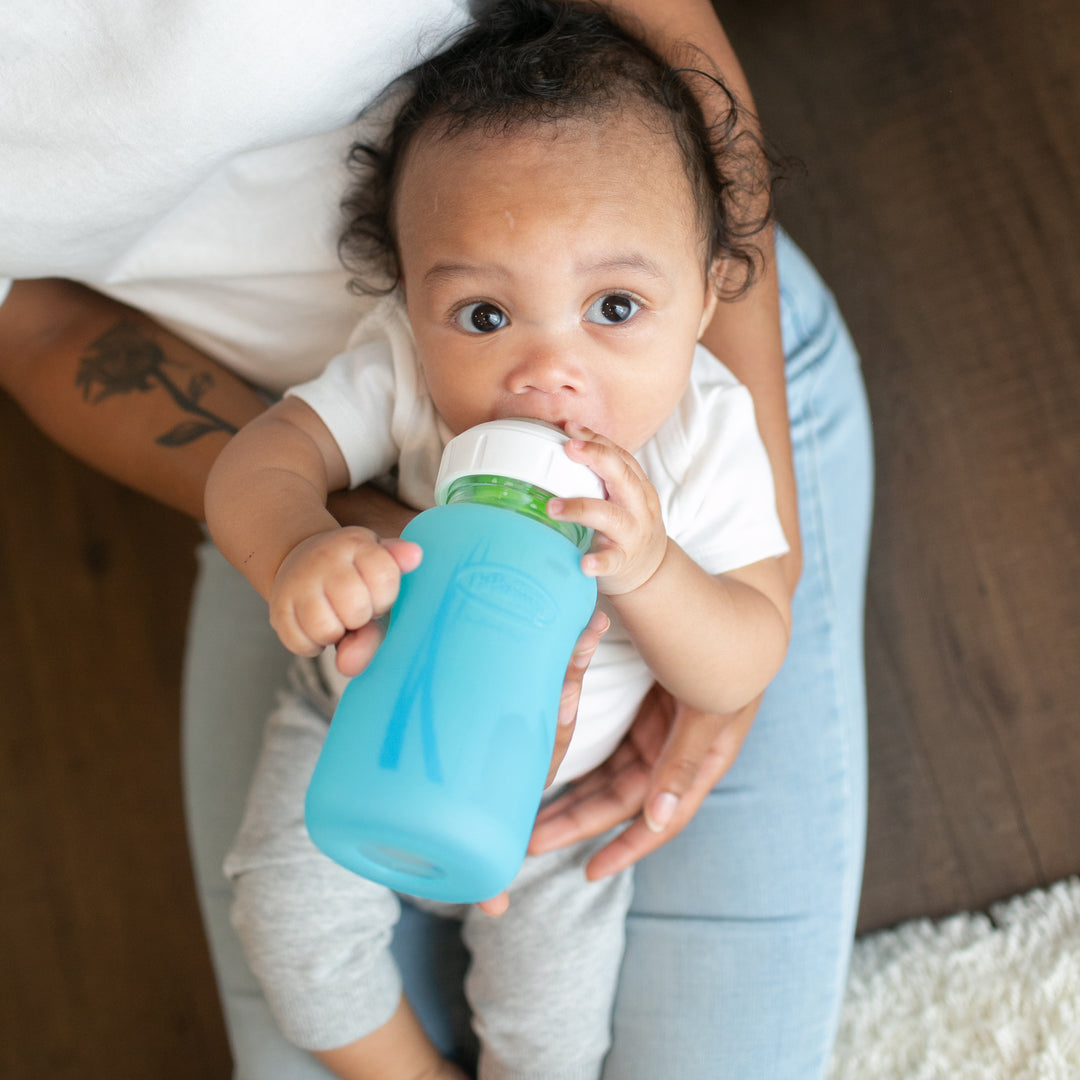 A baby in a white shirt and gray pants sits on an adult's lap, holding a blue sippy cup while looking upwards. The adult, wearing light blue jeans with a tattooed arm visible, watches over the child, who seems ready to switch to Dr. Brown's Natural Flow® Options+™ Wide-Neck Glass Bottle Silicone Sleeves in the future.