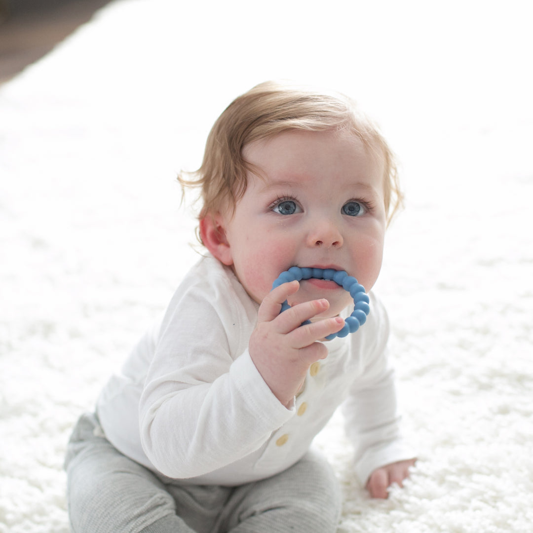 A baby with light brown hair, dressed in a white shirt and gray pants, sits on a fluffy white rug. The child holds Dr. Brown’s™ Flexees™ Beaded Teether Rings to their mouth, finding relief from teething pain as bright sunlight bathes the scene.