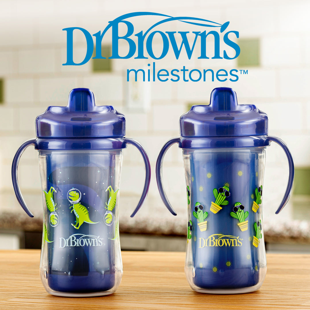 Two Dr. Brown’s Milestones™ Insulated Hard Spout Sippy Cups from the 2-pack set, featuring blue lids and vibrant dinosaur designs, are placed on a table. The kitchen background reveals the brand's logo above these durable cups with bite-resistant spouts.